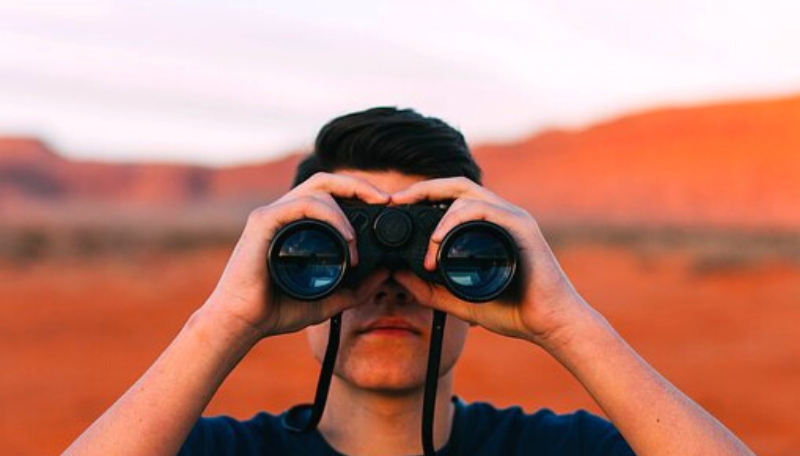 Ein Mensch blickt durch ein Fernglas in die Ferne. Im Hintergrund befinden sich rote Sandberge.
