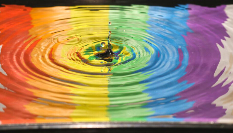Ein Wassertropfen tropft in eine Wasserpfütze, in der sich eine Regenbogenflagge spiegelt.
