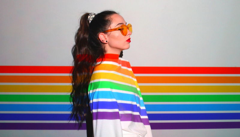 Eine Person mit gelber Sonnebrille steht seitlich und wird von einem regenbogenfarbigen Licht angestrahlt.