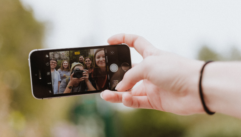 Eine Hand hält ein Smartphone, mit dem gerade ein Bild einer Gruppe junger Menschen gemacht wird.