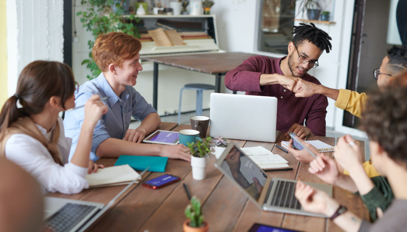 Fünf junge Menschen sitzen gemeinsam an einem Tisch und arbeiten zusammen an Tablets und Laptops.