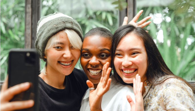 Drei Jugendliche stehen nebeneinander und kommunizieren gemeinsam per Videocall über ein Smartphone. Sie lachen und winken.