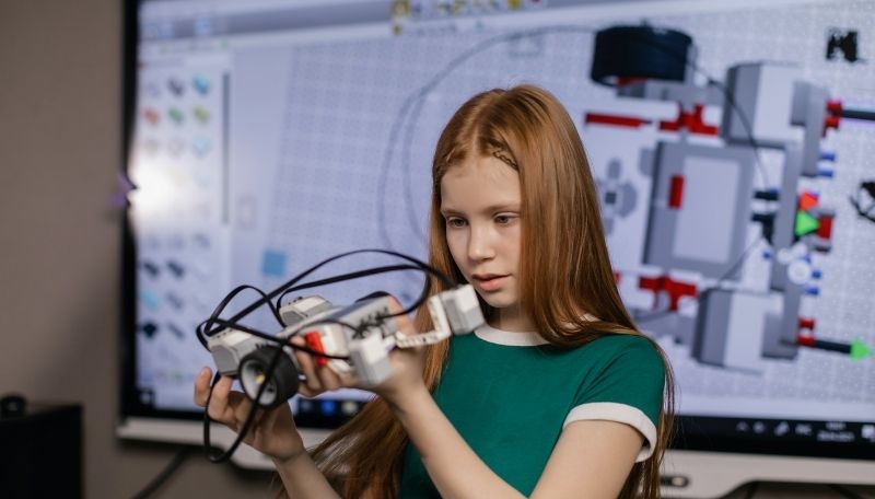 Mädchen bastelt an Fahrzeug aus kleinen Plasik-Bausteinen. Im Hintergrund sind Skizzen auf einem Bildschirm.
